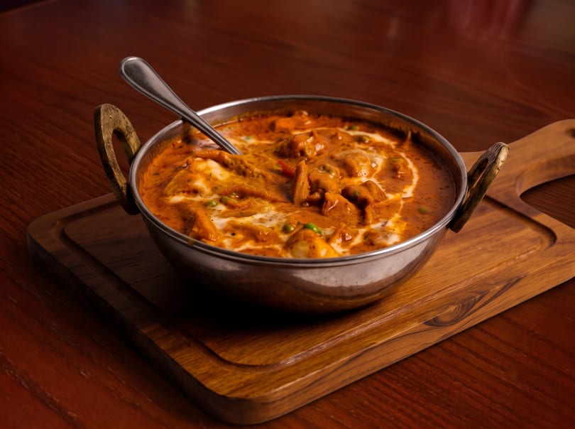A metal karahi bowl filled with creamy butter chicken, garnished with cream swirls, on a wooden board.