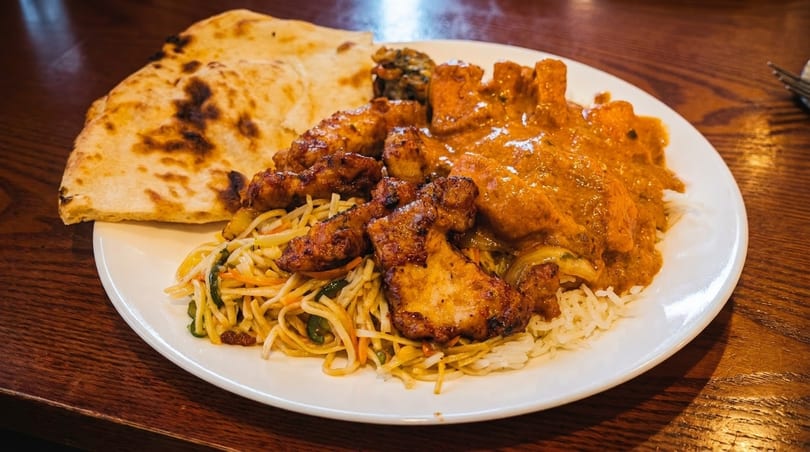 A white plate filled with Indian food, including naan, chicken tikka masala, noodles, and rice.