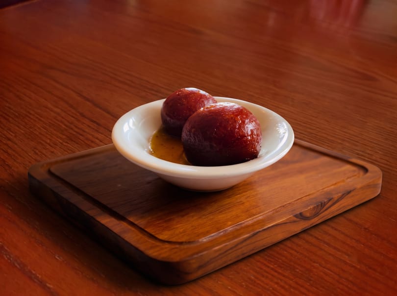 Two gulab jamun in syrup, served in a white bowl on a wooden tray and table.