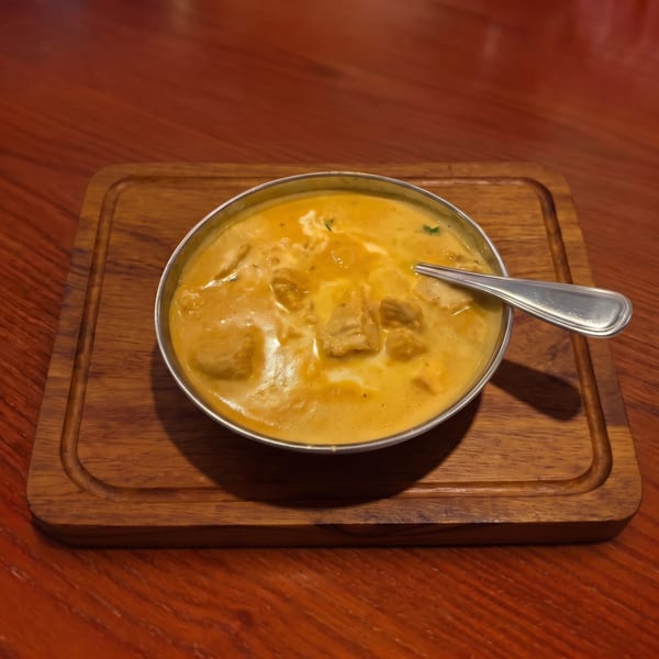 Creamy orange curry with chicken pieces and a spoon, served in a metal bowl on a wooden board.