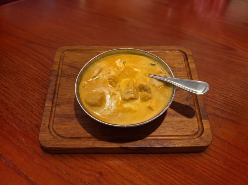 Creamy orange curry with chicken pieces and a spoon, served in a metal bowl on a wooden board.