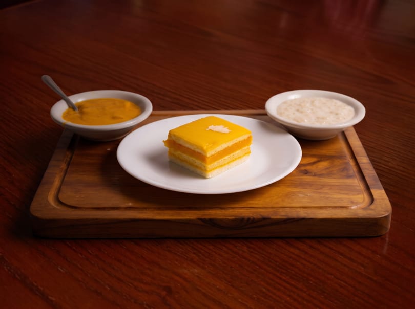 A mango cake slice with two small bowls of sauces on a wooden board.