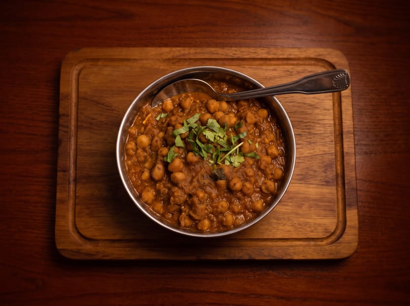 A metal bowl of chana masala, garnished with fresh cilantro, on a wooden serving board.
