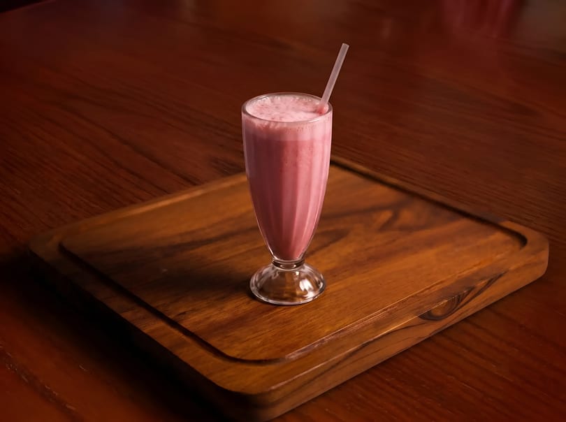 Pink milkshake in a tall glass with a straw, on a wooden serving board.