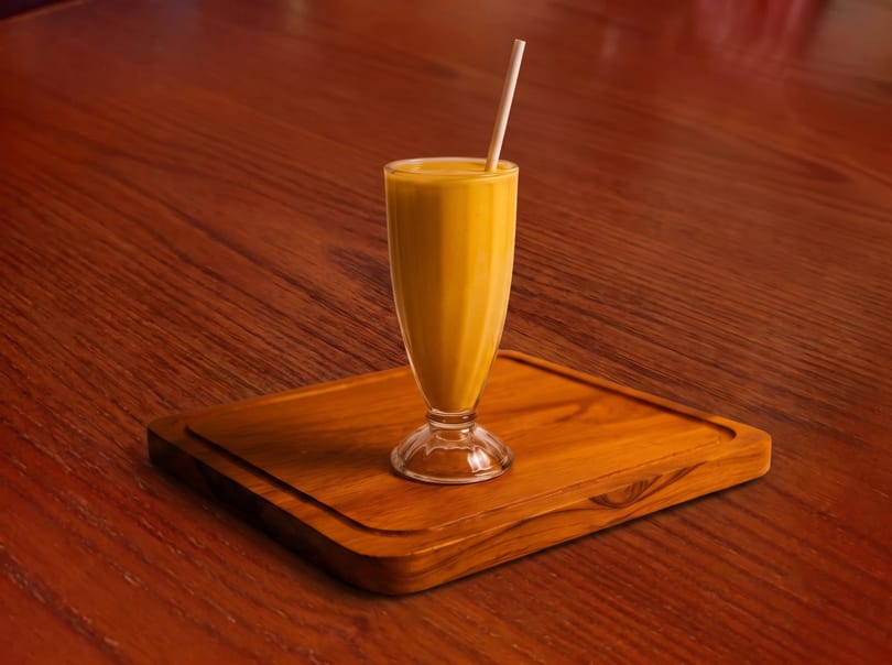 A tall glass of yellow mango lassi with a straw, on a wooden tray and table.