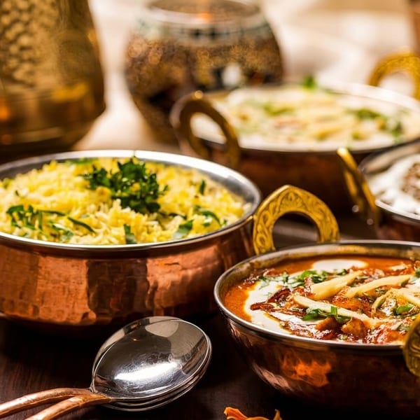 Four copper bowls filled with various Indian dishes, including rice and curries, on a dark wooden table.