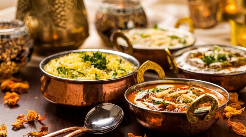 Four copper bowls filled with Indian dishes, including rice, curries, and sauces, on a dark table.