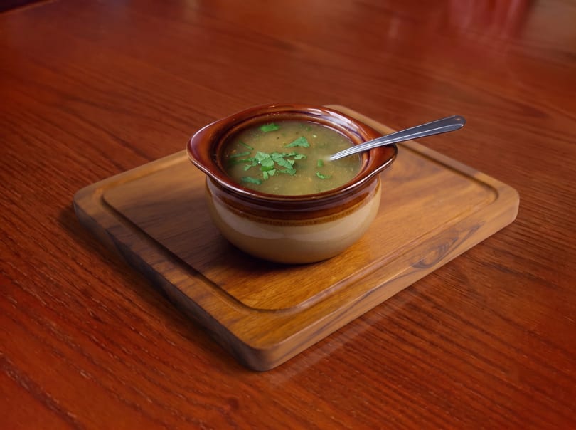 Hearty soup with fresh herbs in a ceramic bowl on a wooden board and table.
