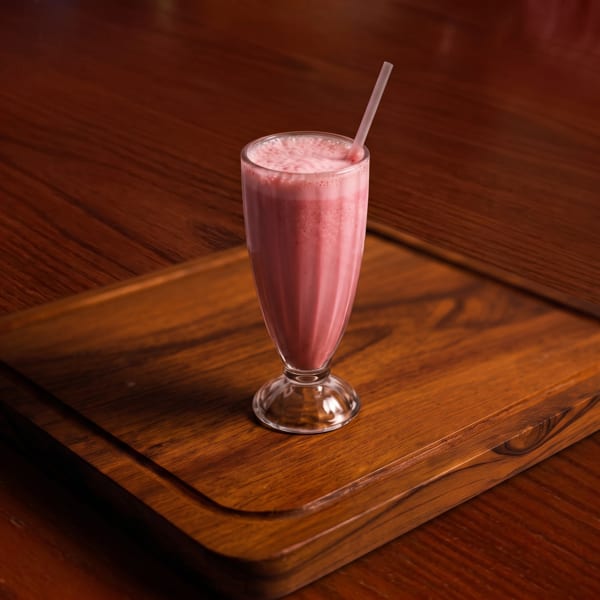 Pink milkshake with a straw in a tall glass, on a wooden serving board.