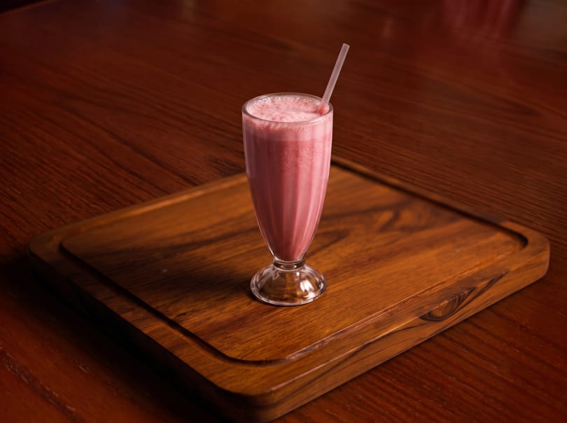 Pink milkshake with a straw in a tall glass, on a wooden serving board.