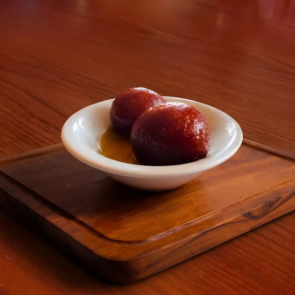 Two gulab jamun in a white bowl with syrup, on a wooden board and table.