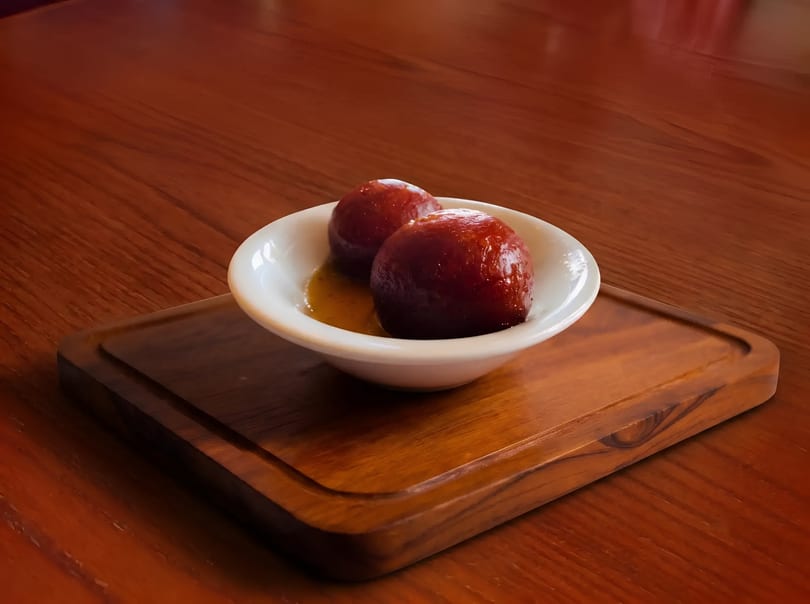 Two gulab jamun in a white bowl with syrup, on a wooden board and table.