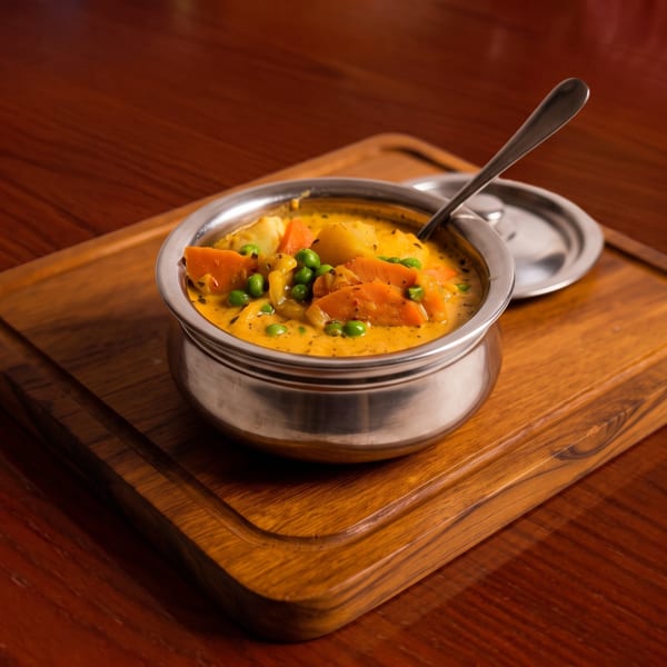 A stainless steel bowl of yellow curry with carrots, peas, and potatoes on a wooden board.