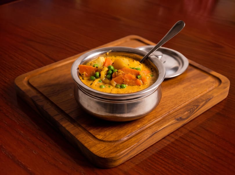 A stainless steel bowl of yellow curry with carrots, peas, and potatoes on a wooden board.