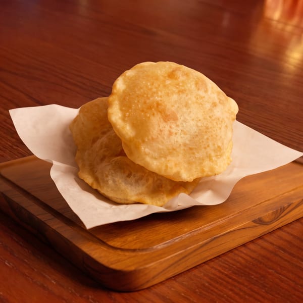 Two golden-brown fried puri breads rest on white parchment paper on a wooden board.