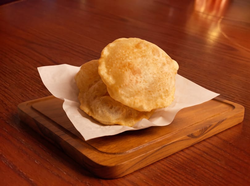 Two golden-brown fried puri breads rest on white parchment paper on a wooden board.